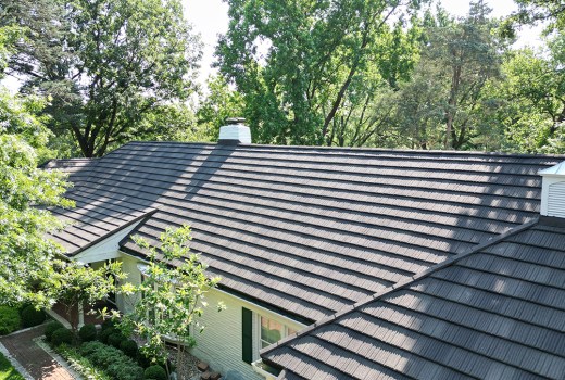 Elevated view of a home featuring a newly installed stone-coated steel roof, showing the textured metal tiles and clean ridge lines. The house is surrounded by mature trees, well-kept landscaping, and bright natural light highlighting the roof’s design and durability.