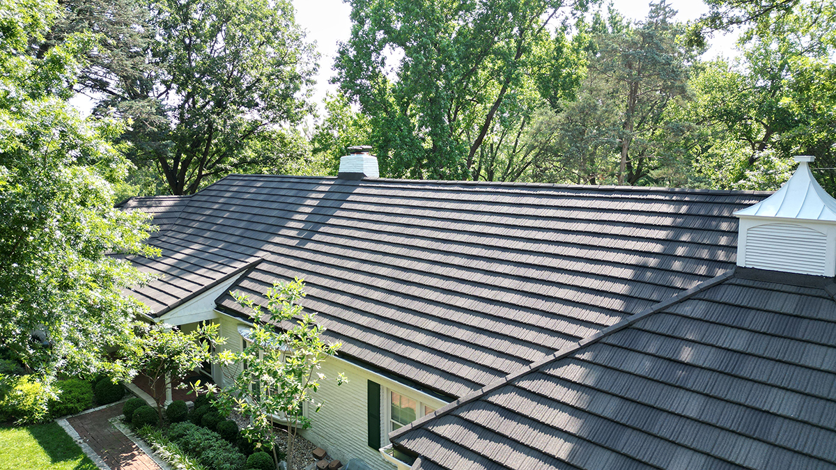 Elevated view of a home featuring a newly installed stone-coated steel roof, showing the textured metal tiles and clean ridge lines. The house is surrounded by mature trees, well-kept landscaping, and bright natural light highlighting the roof’s design and durability.