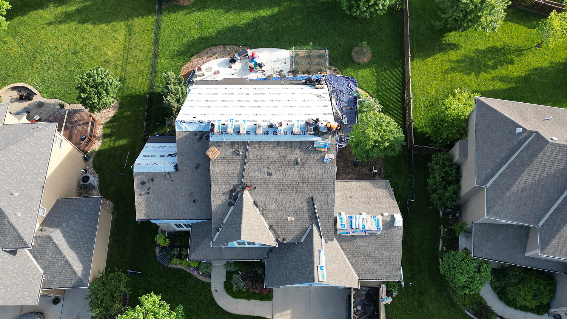 Aerial drone view of a residential roofing project in progress, showing workers installing underlayment and shingles on a large multi-section home. Roofing materials are stacked along the ridges, and tools, tarps, and equipment are visible around the work area. The surrounding homes, lawns, and landscaping create a suburban neighborhood backdrop.